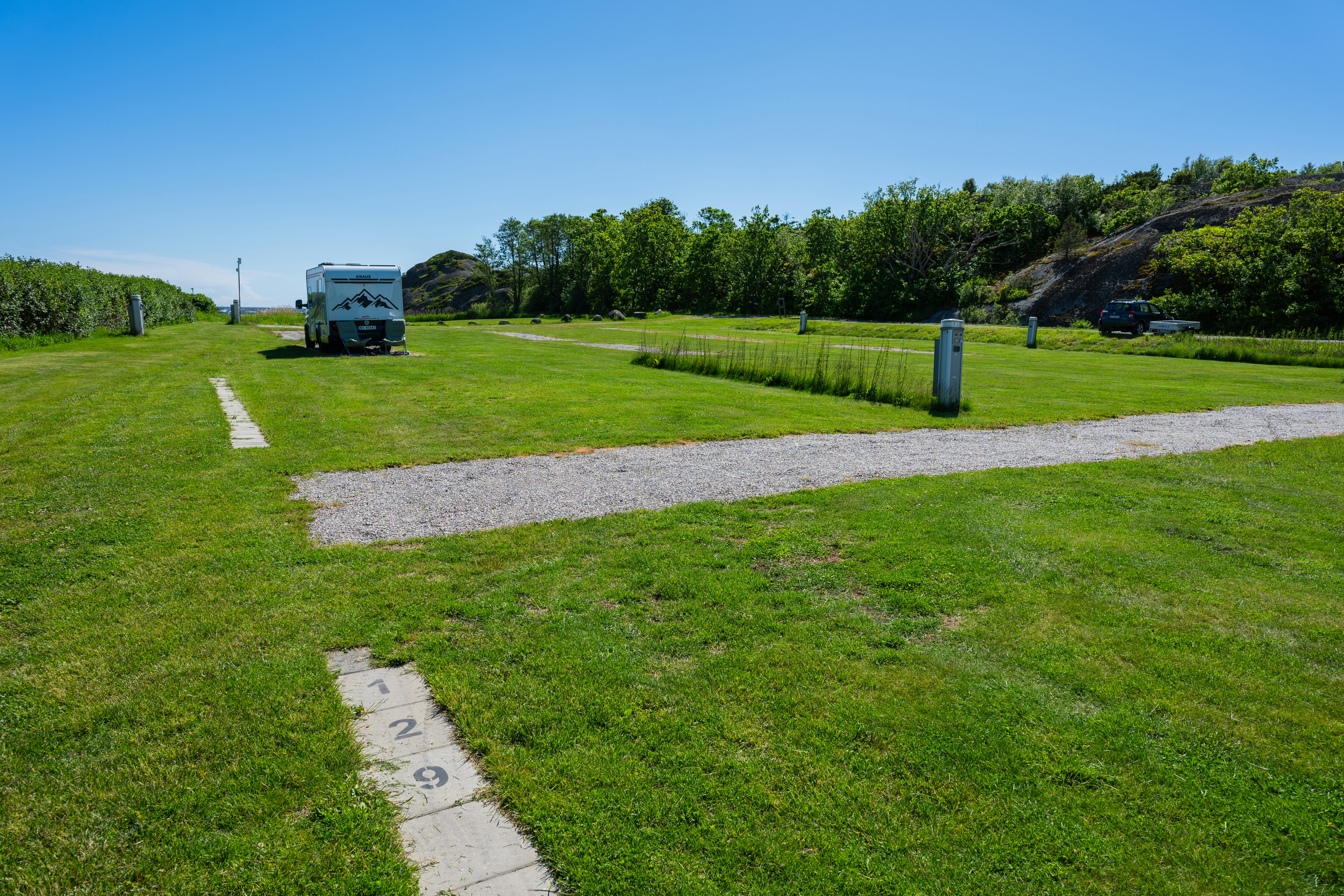 Wohnmobil auf einem Stellplatz auf grünem Rasen, mit Betonwegen, Bäumen und Hecken im Hintergrund.