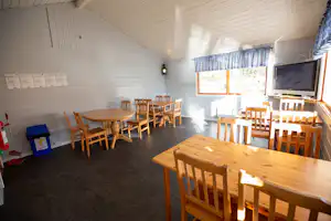 Innenraum eines Campinggebäudes mit Holztischen und Stühlen, TV, Fenster und Sitzbereich an der Wand.