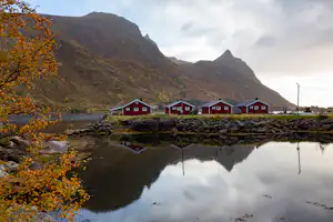 Rote Campinghäuser am See mit Steindamm, Spiegelung im Wasser und Bergen im Hintergrund.