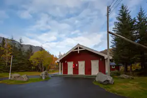 Rotes Campinggebäude mit drei Garagentoren, Einfahrt und Steinen am Rand, umgeben von Bäumen und Hügeln im Hintergrund.