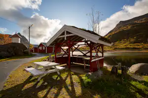 Holz-Pavillon am Seeufer mit Picknicktisch, Weg und roten Gebäuden im Hintergrund, umgeben von Bergen und Bäumen.