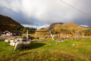 Campingplatz mit grünem Rasen, Spielgeräten und Steinen im Vordergrund, daneben ein Gebäude und Berge im Hintergrund.