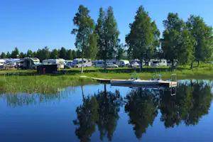 Campingplatz am See mit Wohnwagen und Stellplätzen am Ufer, im Vordergrund ruhiges Wasser und Spiegelung der Bäume.