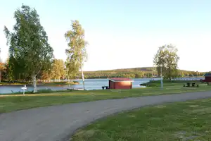 Asphaltweg auf einem Campingplatz am See, mit Rasen, Bäumen und kleinen Gebäuden am Ufer.