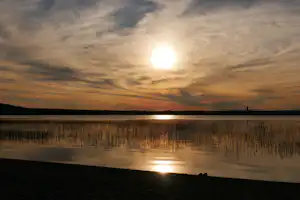Sonnenuntergang über einem See mit Schilf am Ufer und Spiegelung im Wasser, im Hintergrund ein dunkler Horizont.