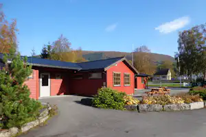 Rote Holz-Hütte mit Eingangstür, großen Fenstern und Terrasse, davor Asphaltweg und Picknicktisch, im Hintergrund bewaldeter Hügel.