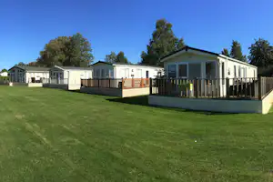 Reihe von Mobilheimen mit Holzterrasse und Zäunen, umgeben von Rasen und Bäumen vor blauem Himmel.