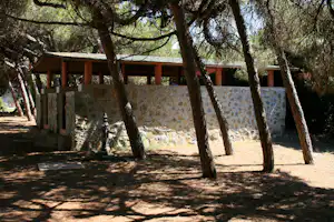 Unterstand am Campingplatz zwischen hohen Bäumen, mit Steinmauer und Holzüberdachung im Hintergrund.