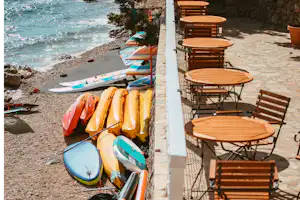 Surfboards am Kiesstrand neben einer Terrasse mit Holztischen und Stühlen, im Hintergrund das Meer.