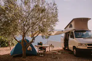 Camping-Van und Zelt auf einem Stellplatz, mit Tisch und Stühlen am Boden, neben einem Baum.