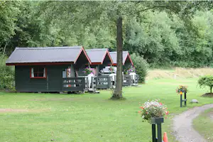 Campinghaus mit Terrasse, Gartenmöbeln und Blumenbeeten vor grünen Bäumen und Rasenfläche.