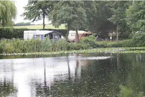 Campingplatz am Fluss mit Wohnwagen und Zelten, umgeben von Bäumen und Schilf am Ufer.