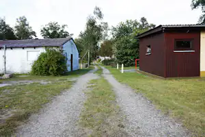 Campingweg zwischen zwei Gebäuden, links ein weißes Haus mit Dach, rechts ein braunes Gebäude, Rasen und Kies.