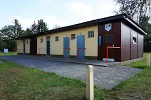 Campinggebäude mit mehreren Eingängen, roten Stufen und gepflastertem Weg, davor Rasen und Bäume am Rand.