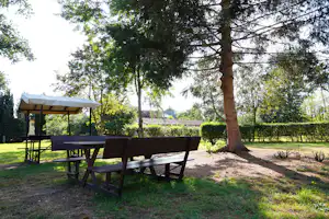 Camping-Tisch mit Bänken unter Sonnenschirm auf einer Wiese, umgeben von Hecken und Bäumen.