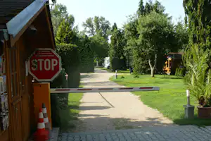 Campingplatz-Weg zwischen Bäumen, links ein Holzgebäude mit Stop-Schild, im Hintergrund Wohnwagen und ein kleines Camp-Gebäude.