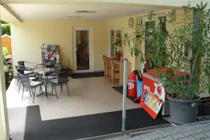 Terrasse am Camping-Eingang mit Sitzgruppe, Tisch, Pflanzenkübeln und Infomaterial an der Wand, überdacht.