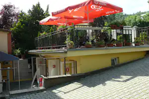 Terrasse mit roten Sonnenschirmen, Holzgartenstühlen und Pflanzkübeln am Campinggebäude, dahinter Bäume und Hecken.