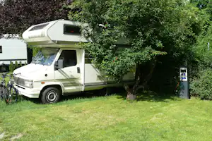 Wohnmobil mit Vorzelt am Campingplatz, neben einem Baum und Hecke, auf grünem Gras mit Stromsäule.