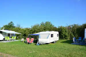 Wohnwagen und Zelt auf einer grünen Campingwiese, daneben Stühle und Sonnenschirm, im Hintergrund Hecken und Bäume.