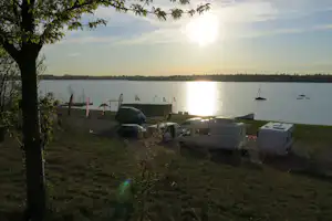 Campingplatz am See mit Wohnwagen und Grill am Ufer, im Hintergrund Boote und die untergehende Sonne.