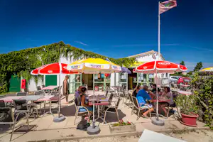 Terrasse eines Campingplatzes mit Tischen, Stühlen und roten Sonnenschirmen, im Hintergrund Gebäude und grüne Hügel.