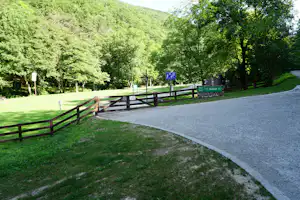 Asphaltierter Campingweg mit Holzgeländern, grüner Wiese und Bäumen am Hang, im Hintergrund ein Wald und Schilder.