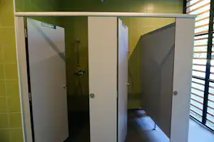 Zwei Duschkabinen mit grauen Türen, Duschkopf und Schlauch, gelb geflieste Wände und Fenster mit Holzlamellen.