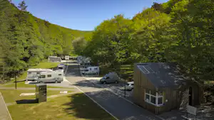 Wohnmobile auf einem Campingplatzweg im Grünen, umgeben von Bäumen und Hügeln, nahe einem Holzgebäude mit Eingang.