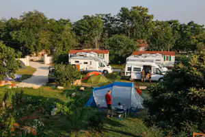 Campingplatz mit Wohnmobilen und Zelten auf dem Stellplatz, gepflegter Rasen, Bäume und Hecken im Hintergrund.