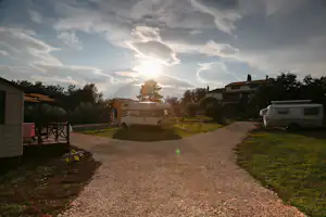 Wohnmobil auf einem Campingplatz-Stellplatz, Kiesweg und Rasenfläche, im Hintergrund Wohnhaus und Bäume bei Sonnenlicht.