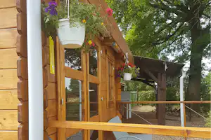 Holzhütte mit Veranda, hängenden Blumenkästen und weißen Pflanzkübeln, im Hintergrund Bäume und Gartenweg.