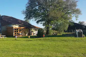 Campinghaus mit Terrasse, Garten und Rasenfläche, umgeben von Bäumen und Hecken, im Hintergrund weitere Gebäude.