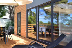 Holzterrasse am Adria-Mobil mit großen Schiebetüren, Tisch und Stühlen sowie Sonnenliegen unter beigen Markisen.