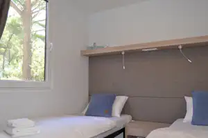 Bett mit weißen Kissen und blauen Kissen in einer Campingunterkunft, großes Fenster mit Blick auf Bäume.