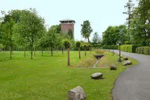Grüner Campingweg mit Hecken, Steinen und Bäumen, im Hintergrund ein Wasserturm und weitere Gebäude.