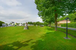 Grünes Campinggelände mit Rasen, großen Bäumen und Wohnwagen am Rand, daneben ein Weg und ein kleines Boot.