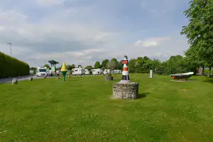 Campingwiese mit Wohnwagen, Boot am Rand und einem Steinsockel mit Laterne, umgeben von Bäumen und gepflegtem Rasen.