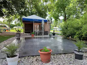 Holzhütte mit blauem Dach auf dem Campingplatz, davor eine Stein-Terrasse mit vielen Blumentöpfen und Wegen im Grünen.