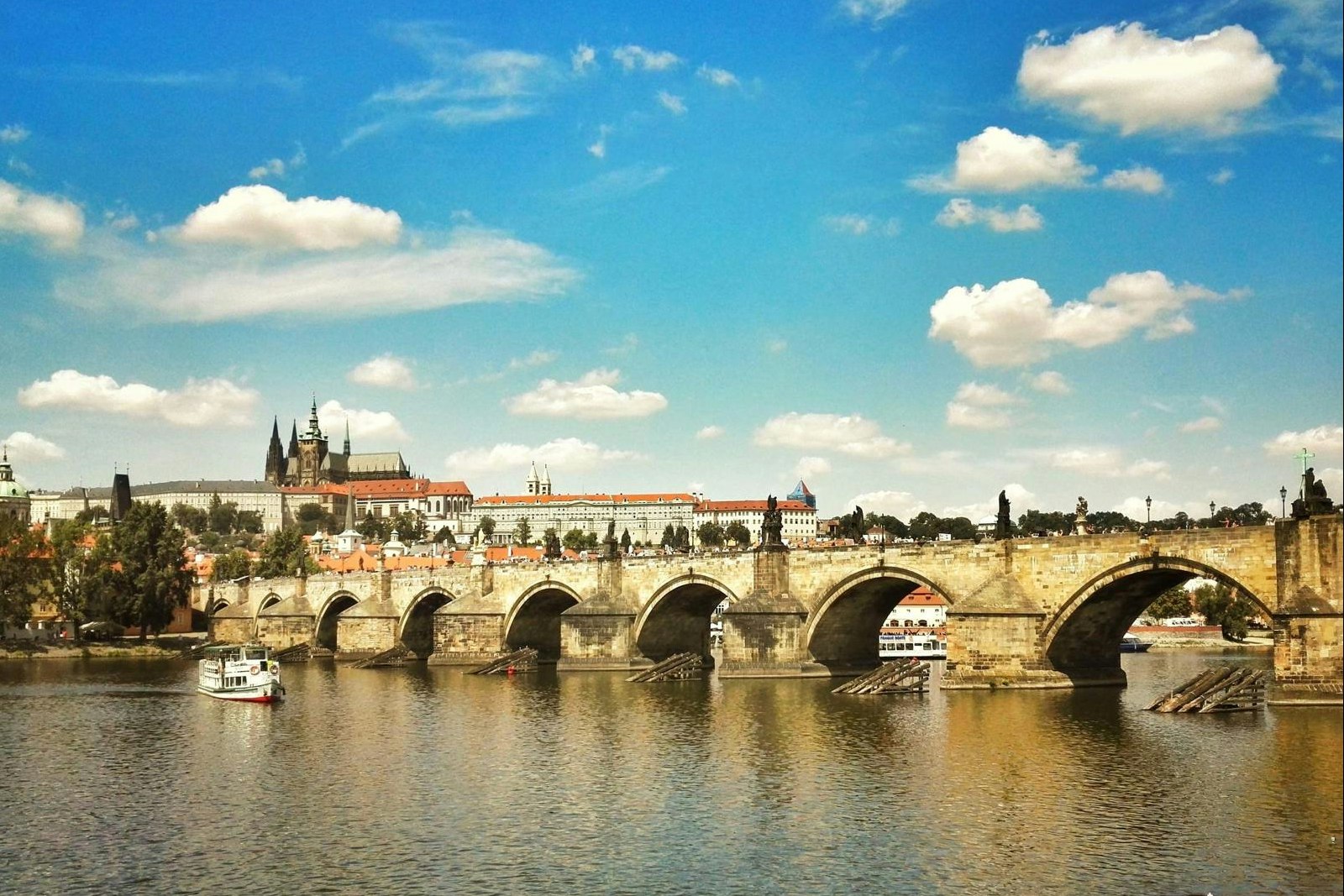 Steinbrücke über dem Fluss mit mehreren Bögen, Boote im Wasser und historische Gebäude am Ufer unter blauem Himmel.
