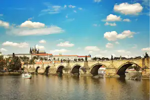 Steinbrücke über dem Fluss mit mehreren Bögen, Boote im Wasser und historische Gebäude am Ufer unter blauem Himmel.