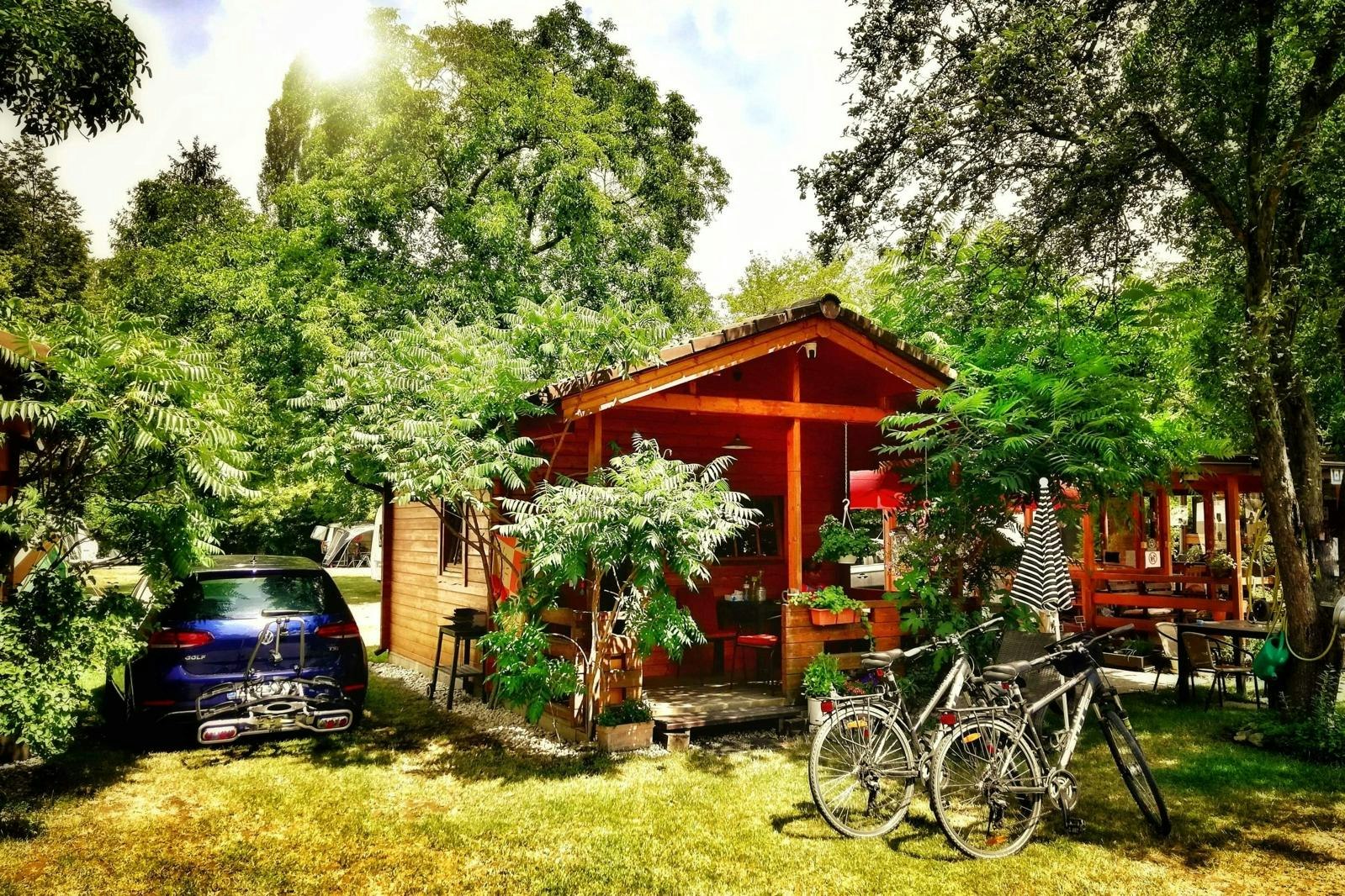 Holzunterstand mit Sitzbereich im Garten, Fahrräder davor, Auto am Rand, umgeben von Bäumen und Grünpflanzen.