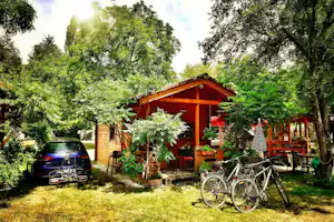 Holzunterstand mit Sitzbereich im Garten, Fahrräder davor, Auto am Rand, umgeben von Bäumen und Grünpflanzen.