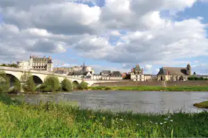 Fluss mit Gras am Ufer, Steinbrücke und Schlossanlage mit Türmen und Kirchengebäude im Hintergrund.
