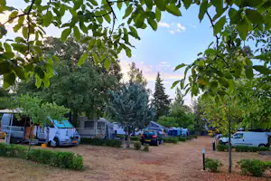 Wohnwagen und Autos auf einem Campingplatz, umgeben von Bäumen und Sträuchern, mit Kiesweg und Stellplätzen.