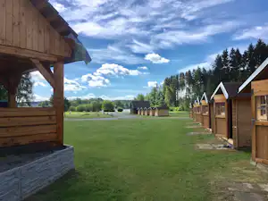 Holzhütten auf einer grünen Campinganlage, Blick zwischen Holzfassaden und Wegen, im Hintergrund hohe Bäume und Himmel mit Wolken.