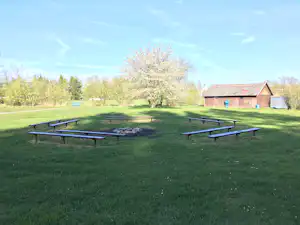 Grüne Wiese mit mehreren Picknicktischen und Grillstelle vor einem Holzgebäude, im Hintergrund Bäume und ein kleiner Teich.