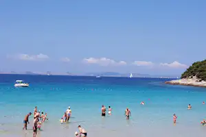 Badebucht am Meer mit Schwimmern im türkisfarbenen Wasser, Segelboote in der Ferne und Felsufer mit Bäumen.