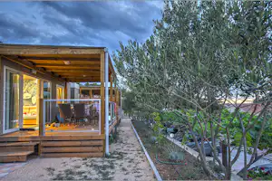 Mobilheim mit offener Terrasse, Holzdach und Sitzgruppe; im Vordergrund Kiesweg und Olivenbaum am Rand des Stellplatzes.