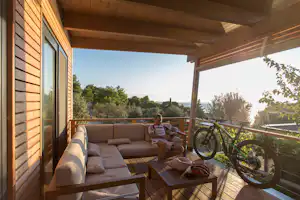 Holzterrasse am Campingunterkunft mit Sofa, Tisch und Fahrrad, Blick ins Grüne und auf das Meer bei Sonnenlicht.